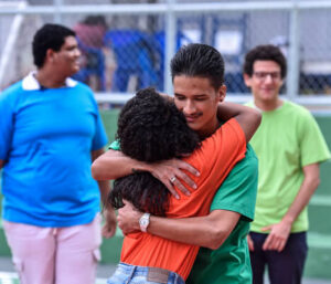 No centro dois jovens se abraçando e mais dois jovens atrás.