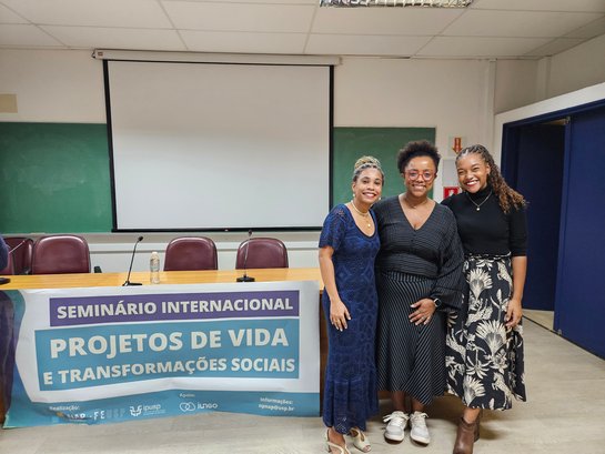 Mesa com cartaz, telão desligado ao fundo. No lado direito da mesa, três mulheres negras, de pé, sorrindo para a foto.