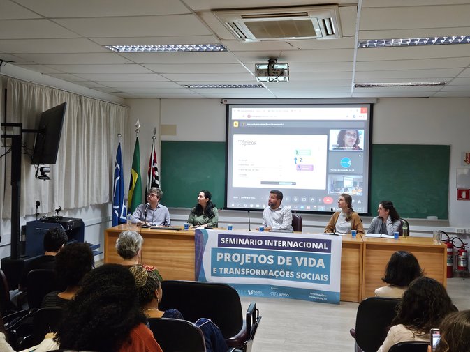 Palestrantes sentados atrás de uma mesa, com um telão atrás, em auditório da USP. Plateia de frente, sentada.