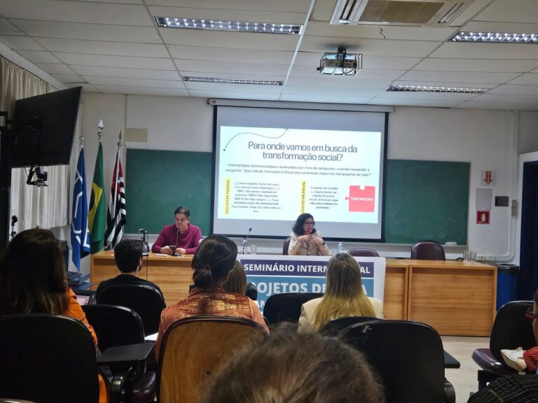 Palestrantes em um auditório da USP, em um palco, sentados, com uma mesa a frente. Um telão atrás dos palestrantes e a plateia a frente.