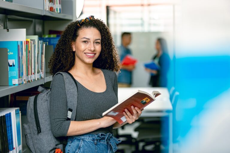 Imagem de uma biblioteca, uma mulher de pé próxima a uma estante de livros, de mochila, blusa escura e calça jeans, com um livro aberto nas mãos e sorrindo.