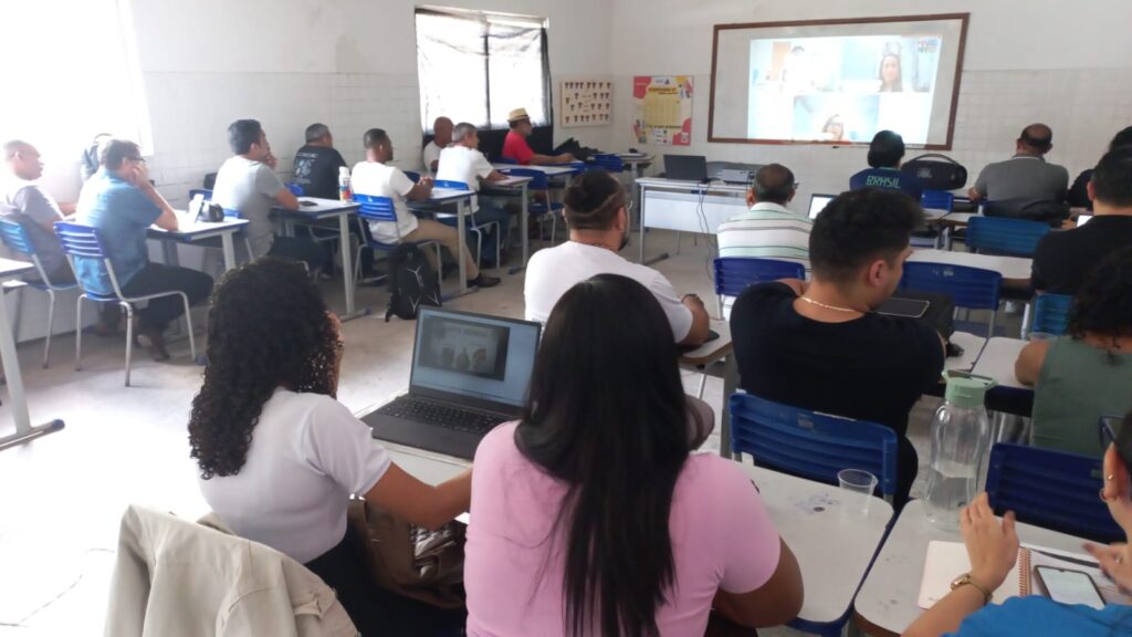 Professores reunidos em sala de aula, assistindo um telão.