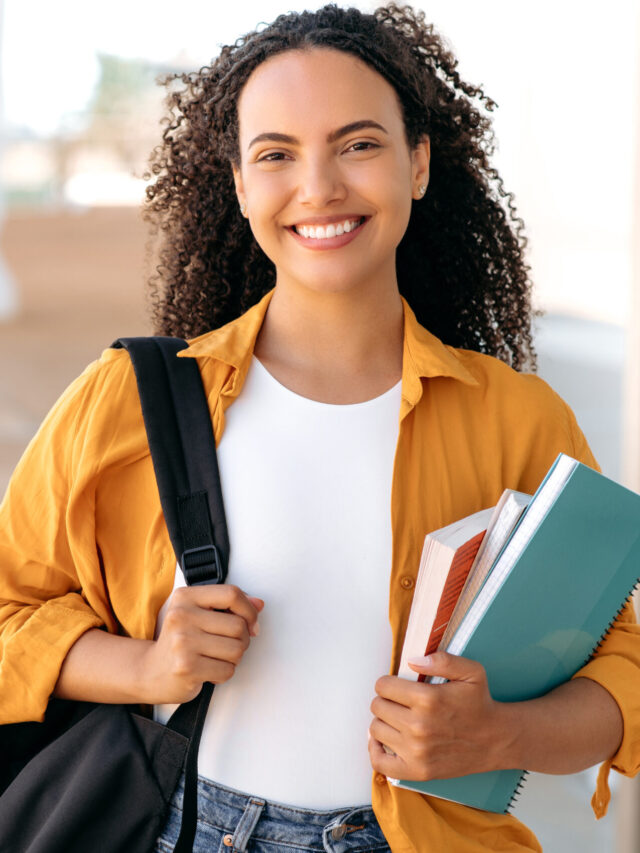 Mulher jovem de cabelo cacheado com mochila e livros na mão sorrindo.