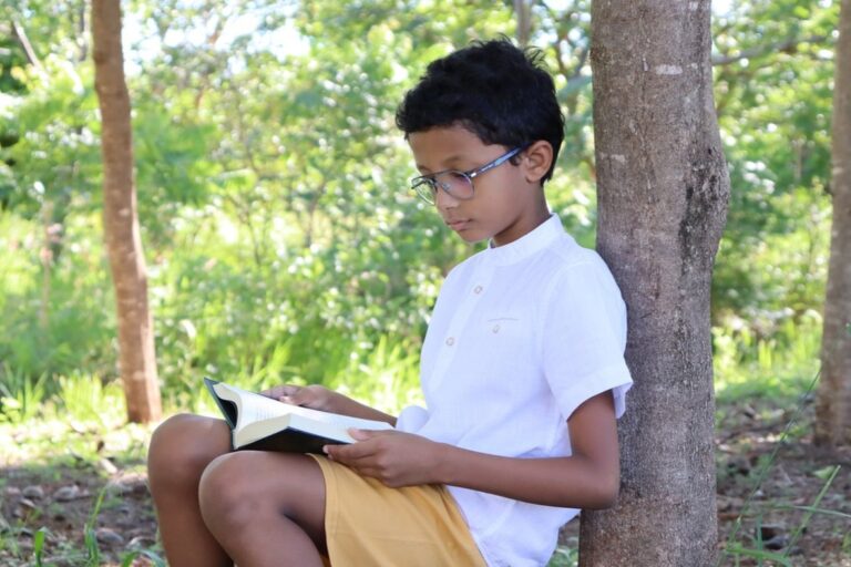 Criança lendo um livro, em local cercado de árvores, durante o dia.